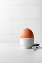 Boiled egg in an egg stand and spoon on a white kitchen background with text space. Light protein...