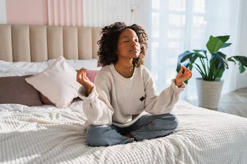 Foto auf Acrylglas Yoga mindfulness meditation. Young healthy African girl practicing yoga at home. Woman sitting in lotus pose on bed meditating smiling relaxing indoor. Girl doing breathing practice. Yoga at home © Юлия Завалишина