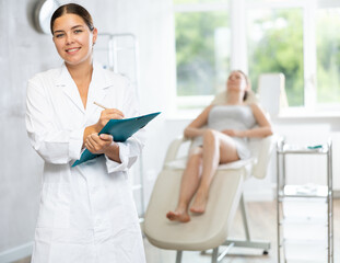 Obraz premium Woman doctor holds folder with papers in hands and enters medical information about client of clinic into form. In background, female visitor is lying in armchair and chatting with specialist