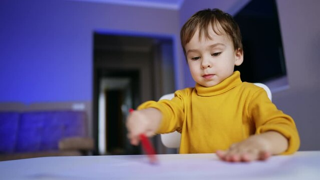 Beautiful toddler boy sits at desk drawing. Cheerful baby raises hands smiling happily. Low angle view. Close up.