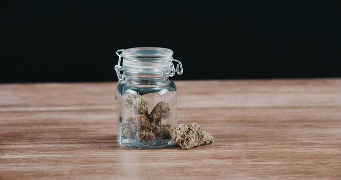 Weed, jar and marijuana in glass on table with dry herbs, drugs or cannabis flowers in dark background of studio. Bottle, container and storage of legal medical pot, cbd and thc product on desk