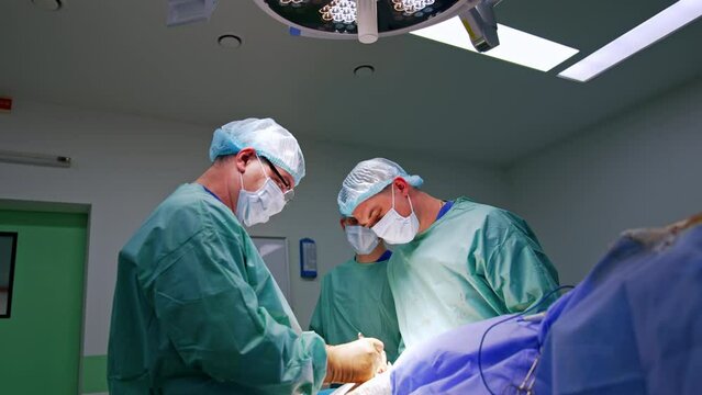 Doctors standing on both sides of a patient looking focused at operated area. Main surgeon applies metal instruments.