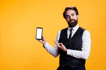 Professional employee advertises white screen on mobile phone, showcasing empty isolated blank display with copyspace against yellow background. Young man presents blank mockup device.