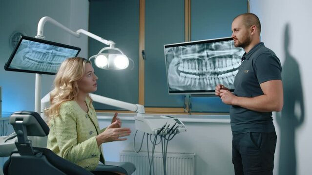 Handsome young athletic male dentist answering questions to beautiful blonde female patient sitting in dentist chair, illuminated fully equipped office. High quality 4k footage