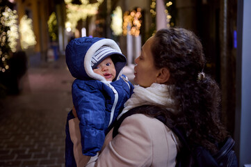 Mother holding her baby boy looking at camera, standing in the street at night, decorated with garlands for Christmas