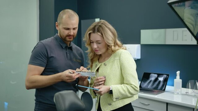 Bald athletic dental expert in T-shirt selecting implants for charming elegant blonde with hand mirror, looking at teeth, standing in modern dentist office. High quality 4k footage