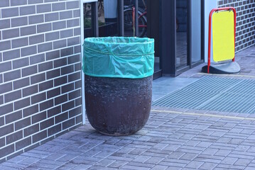 one brown concrete trash can with a green plastic bag stands on a gray sidewalk near a brick wall on the street