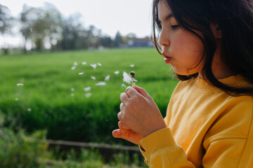 side view of child in yellow top blowing a dandelion outside in a green field