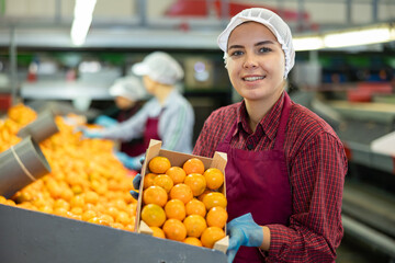 Happy young female worker of citrus sorting factory showing ripe selected mandarin oranges packed...