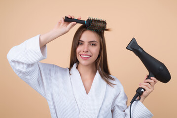 Woman in bathrobe combing hair, drying hair with hairdryer. Portrait of female model with a comb brushing hair. Girl with hairs brush and blow dryer. Hair care and beauty. Morning routine. - obrazy, fototapety, plakaty