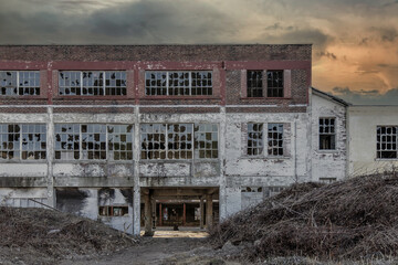 Facade of a derelict former factory building with broken windows, missing roof, piles of debris in foreground, dark sky, nobody