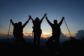 Silhouette of Asian three people standing raised hands with trekking poles and kerosene black lamp on cliff edge on top of rock mountain with at sunset rays over the clouds background,
