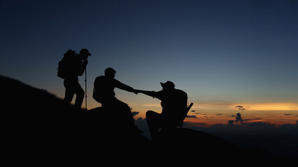 Fototapeta na wymiar Silhouette of Asian teamwork hikers climbing up mountain cliff and one of them giving helping hand with friend at sunset, people helping, team work concept.