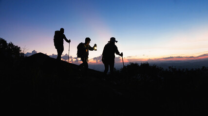 Silhouette of Asian teamwork hikers climbing up mountain cliff and one of them giving helping hand with friend at sunset, people helping, team work concept.