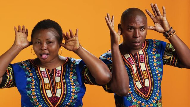 Silly ethnic couple fooling around on camera with comic face, having fun and sticking tongue out in studio. Cheerful people doing funky gesture and enjoying time on set, orange background.