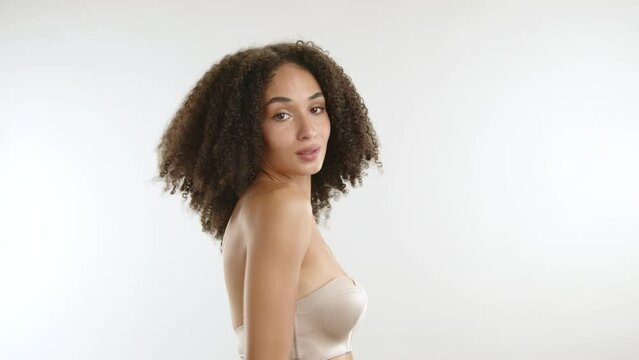 Attractive young woman with african curls, slim silk body wearing strapless bra, hugging herself gently, touching body seductively. White background. High quality 4k footage
