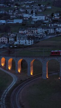 Train on Illuminated Brusio Spiral Viaduct in Switzerland in the Evening. Bernina Railway. Swiss Alps. Aerial View. Orbiting. Vertical Video