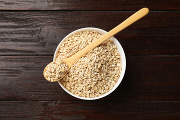 Dry pearl barley in bowl and spoon on wooden table, top view