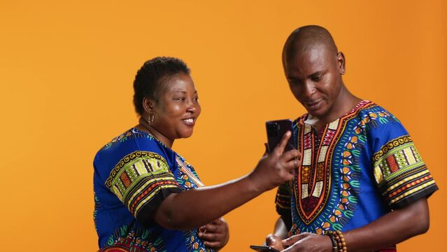 Happy african american people scrolling webpages on smartphone, checking pictures on social media app and having fun with memes. Married couple in traditional attire looking at photos online.
