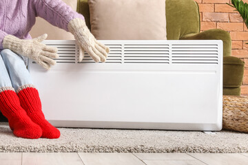 Young woman in warm winter socks and gloves near electric heater. Concept of heating season