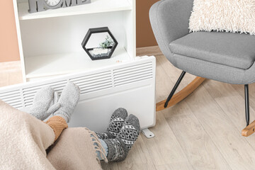 Young woman in winter clothes with warm plaid near electric heater at home. Concept of heating...