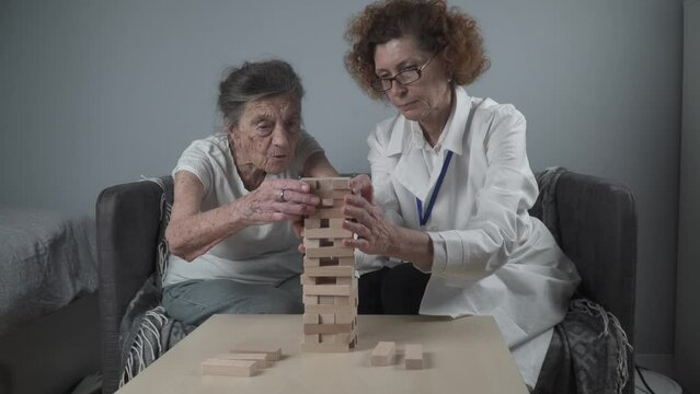 Dementia therapy in playful way, training fingers and fine motor skills, build wooden blocks into tower, playing Jenga. Senior woman 90 years old and doctor playing educational game in nursing home.