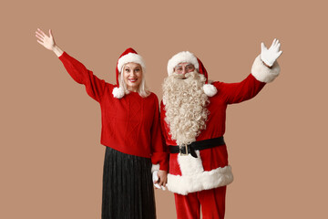 Santa Claus and beautiful mature woman in Christmas hats waving hands on brown background