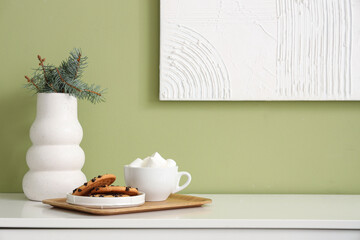 Vase with fir branches, cup of cocoa drink and cookies on table near color wall