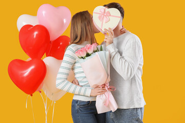 Lovely couple with gift box, bouquet of flowers and heart-shaped balloons on yellow background....