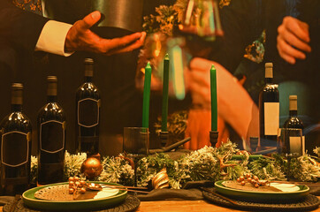 Side view of dark wine bottles, wine glasses, green candles and decorations on a table with human hands in background.