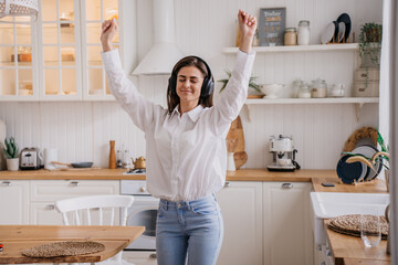 Joyful young woman in a crisp white shirt dances freely in a bright kitchen, headphones on, lost in...