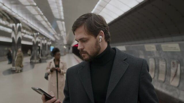 Tilt up medium shot of young adult Caucasian businessman wearing wireless earbuds while waiting for subway train at station