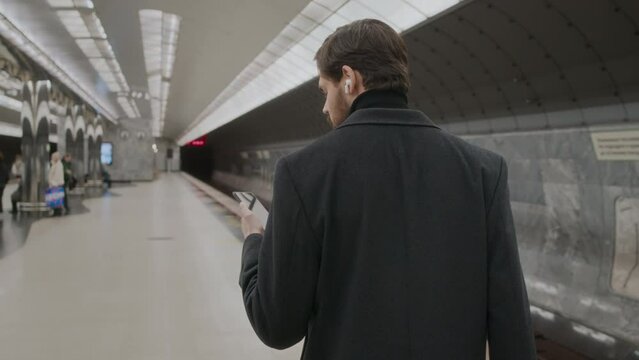 Back view medium shot of Caucasian man in black blazer coat checking his smartphone while going along railroad at subway station, waiting for train