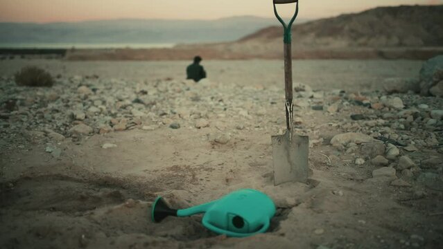 Watering can and shovel on sand with nomad man silhouette in background at sunset