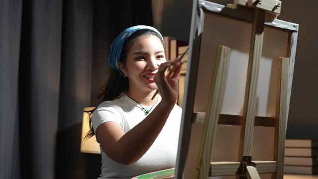 A young Asian female artist and painter paints with a paintbrush with painting on an easel in the art studio with acrylic paints on canvas. Lifestyle hobby ideas.