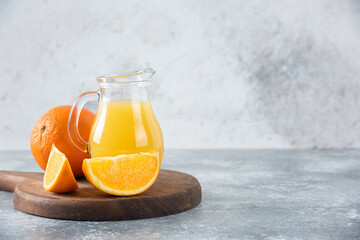 A glass pitcher of fresh juice with whole and sliced orange fruit placed on wooden board