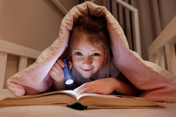 funny preschool girl lying in bed and reading book with flashlight under blanket