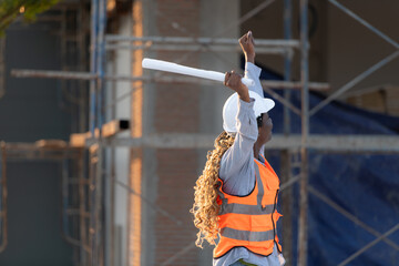 At the building site, a female engineer holds a blueprint in hand and raises her hands in delight.