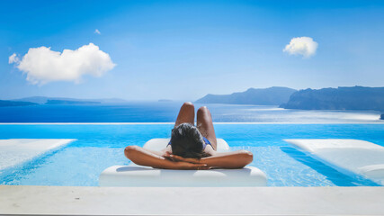 Naklejka premium Young Asian women on vacation at Santorini relaxing in a swimming pool looking out over the Caldera ocean of Santorini, Oia Greece, Greek Island Aegean Cyclades during summer in Europe