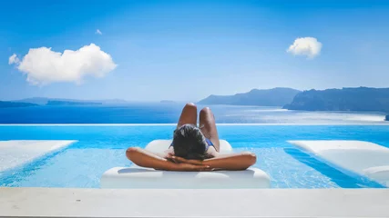 Poster Young Asian women on vacation at Santorini relaxing in a swimming pool looking out over the Caldera ocean of Santorini, Oia Greece, Greek Island Aegean Cyclades during summer in Europe © Fokke Baarssen