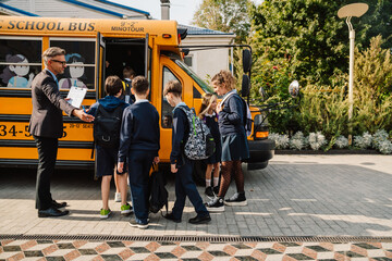 Friendly male teacher holding clipboard and checking off children names while they getting on school bus