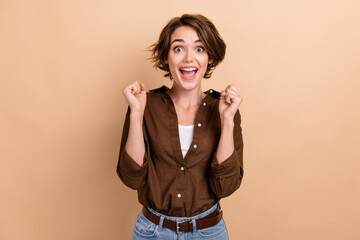 Photo of cute lucky lady wear brown shirt rising fists shouting yeah isolated beige color background
