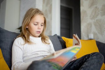 A younger schoolgirl with blonde hair is reading an interesting book, enjoying the silence and comfort of home.