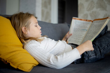 Fototapeta na wymiar Immersed in reading, a blonde younger schoolgirl enjoys a moment of homely comfort on the couch.