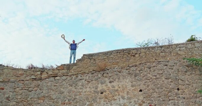 A young adult man wearing a hat and glasses stands on a medieval wild stone wall with his arms raised up. The gesture expresses joy and freedom