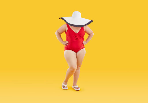 Full length shot of fat, plump, plus size woman model, wearing red swimsuit, beach flip flops and big wide brimmed summer sun hat that hides her face, is posing isolated on yellow color background