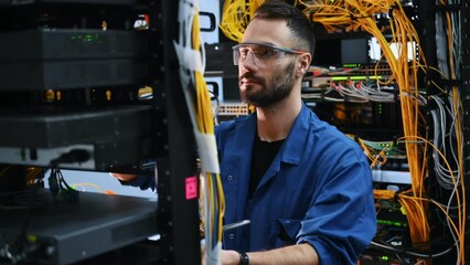 Holding smartphone. Young man is working with internet equipment and wires in server room.