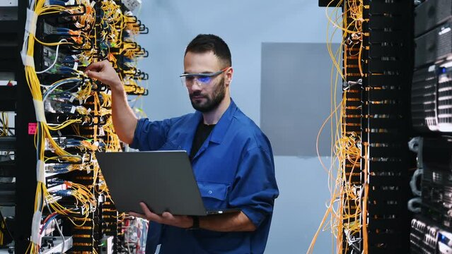 With laptop in hands. Young man is working with internet equipment and wires in server room.