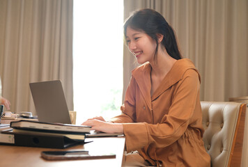 Young attractive Asian woman smiling thinking planning writing in notebook, tablet and laptop working from home, looking at camera at modern office