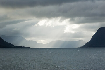 A body of water with mountains and sun striking through the cloudsin the background